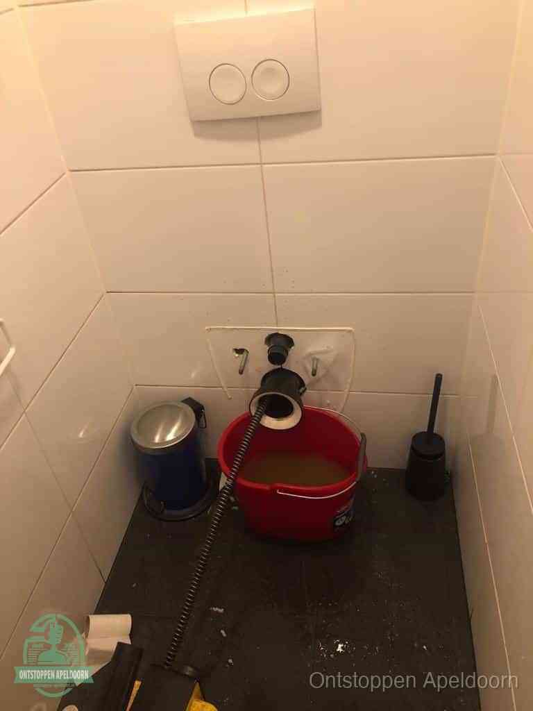 Een ontstopte wand-wc met verwijderde toiletpot waar een ontstoppingsveer in de wanduitgang verdwijnt, het achterhalen waar de verstopping zit in Apeldoorn