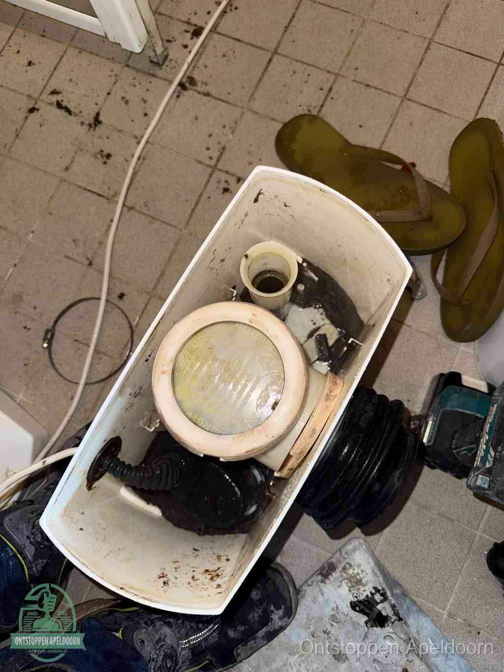 Een gedemonteerde toiletpot op de vloer met ontstoppingsveer en gereedschap ernaast, het verwijderen van een vastzittend voorwerp uit de wc in Apeldoorn